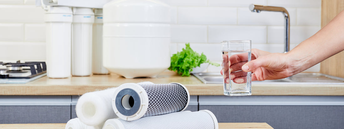 Someone holds a glass of clean water up from a kitchen counter with under-sink drinking water filters sitting on the same counter