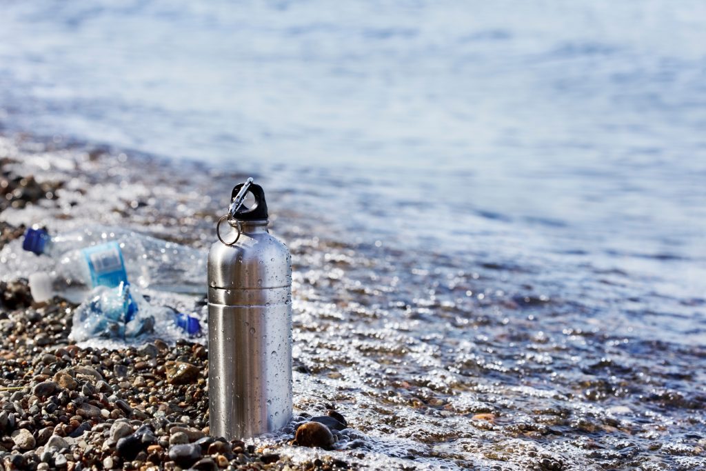 A metal water bottle sitting on a rocky beach with plastic water bottles pulled in by the tide