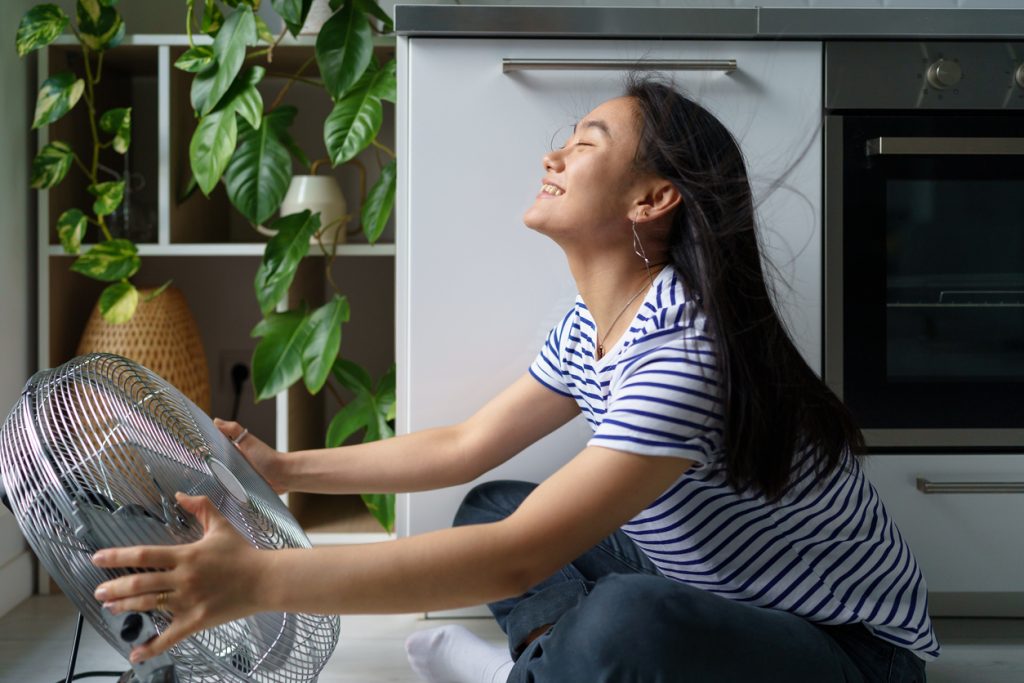 A young Asian woman sits in front of an electric floor fan at home, enjoying the clean air thanks to her UV air purification system